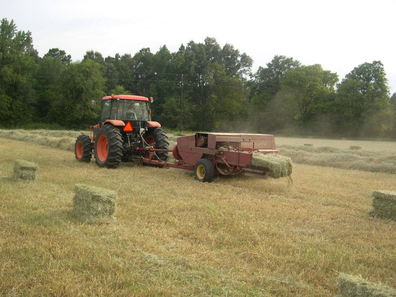 Orange tractor baling hay in a field, pulling a red square baler with dust rising.