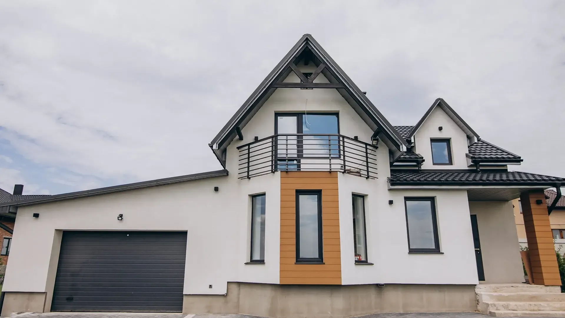 White two-story house with black roof, garage door, and balcony against a cloudy sky.
