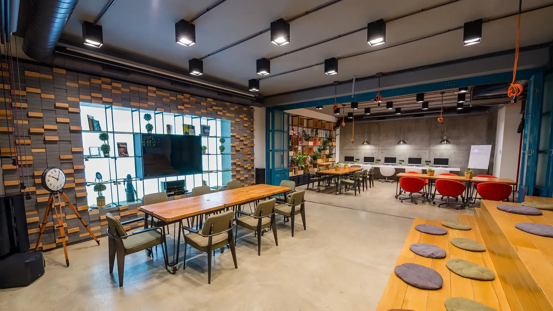 Modern office space with large tables, chairs, and multiple work areas. Exposed ceilings, brick and wooden accents.