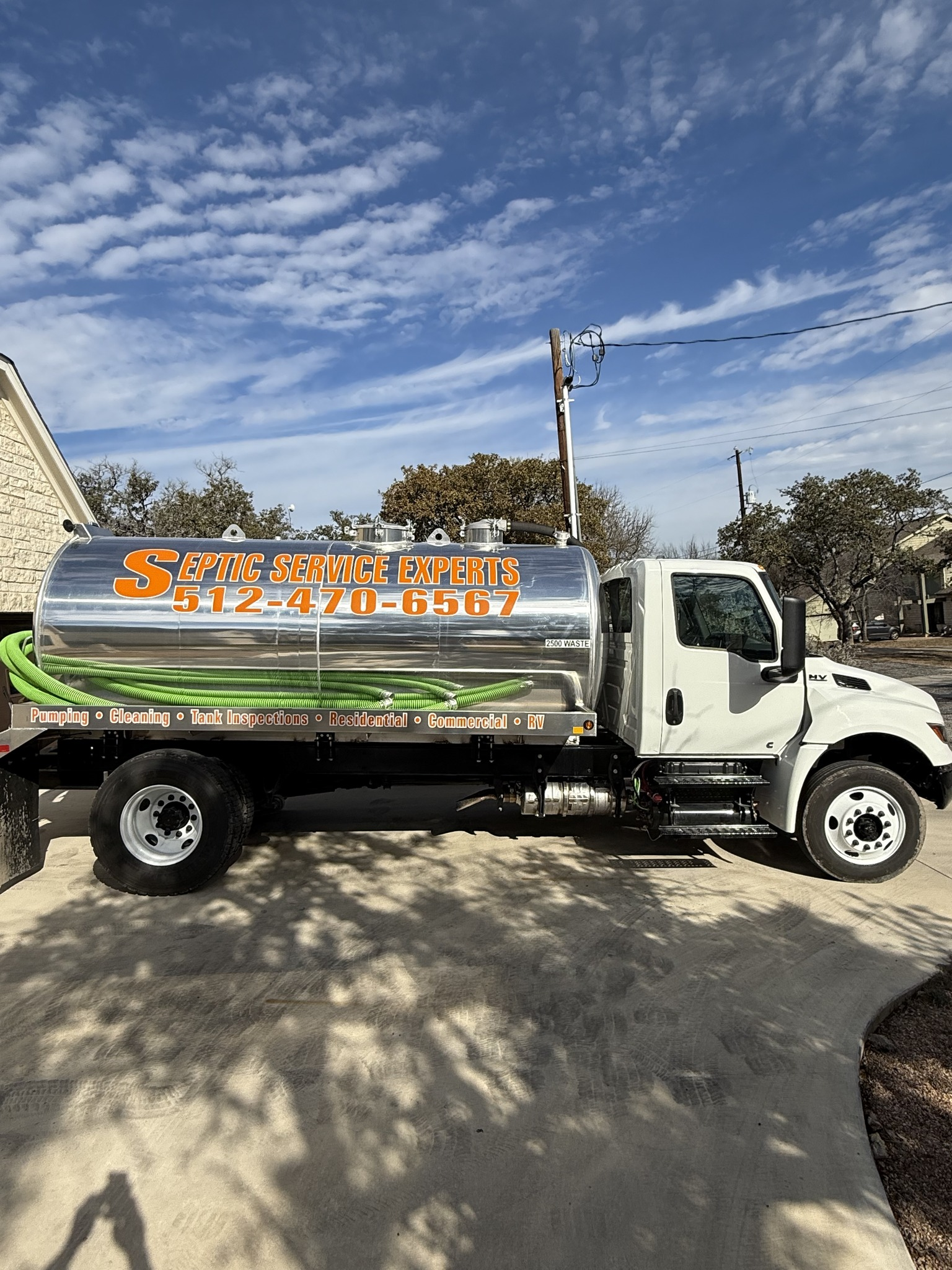 Septic service truck with a silver tank and green hose parked on pavement under a blue sky.