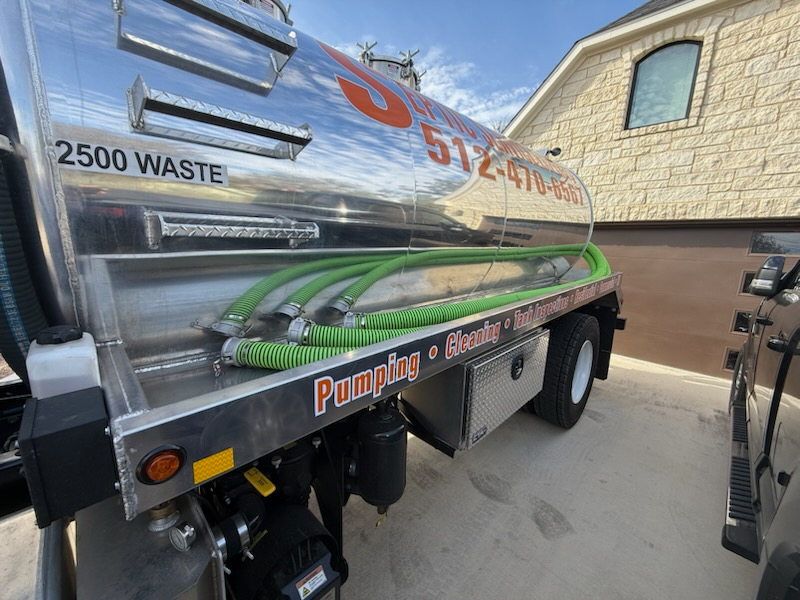 A silver septic tank truck with green hoses parked in front of a tan brick building.