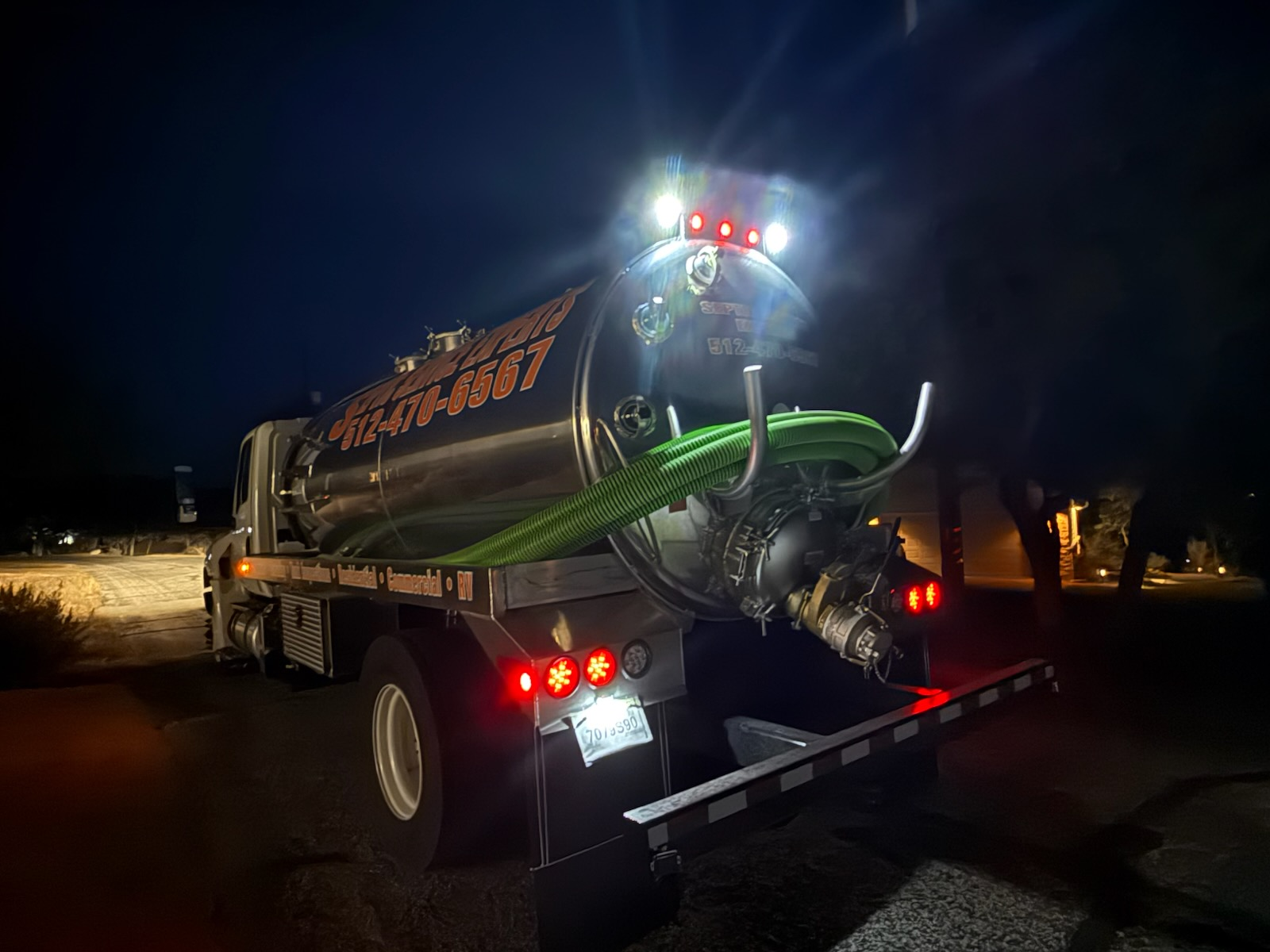 A septic tank pumping truck parked at night with a green hose, with lights on.