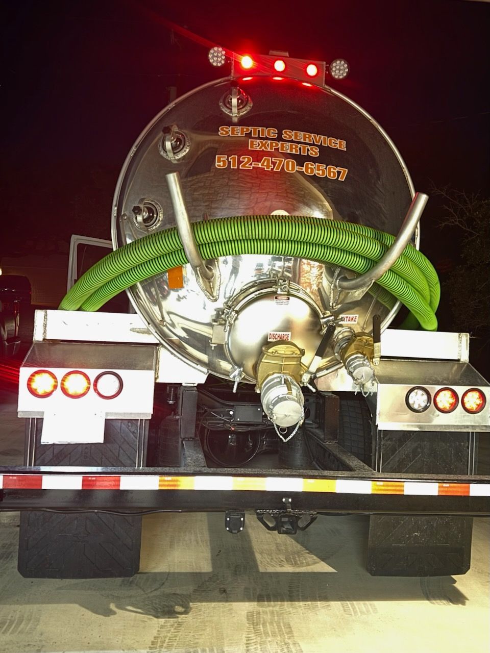 Rear view of a septic truck at night, with a shiny tank, green hoses, and red lights illuminated.