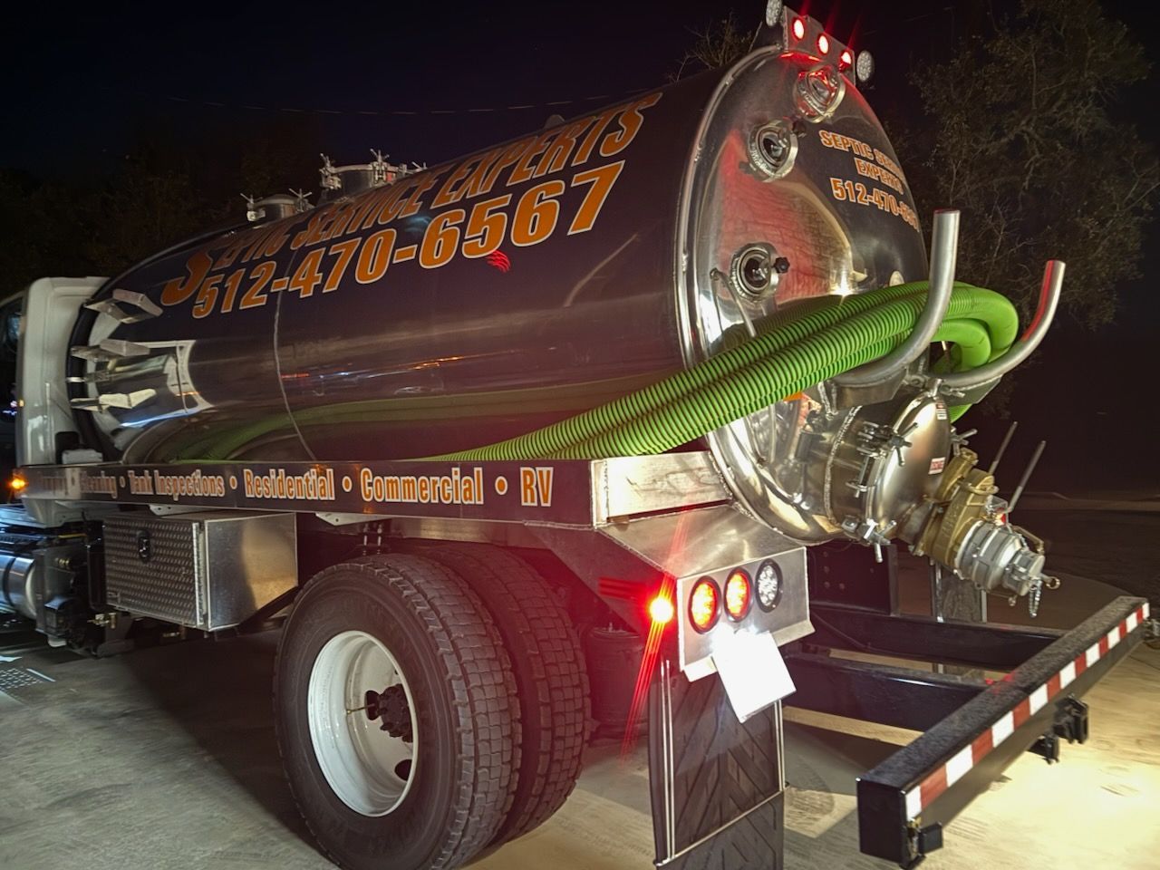A septic tank truck with a shiny silver tank, green hose, and flashing red lights at night.