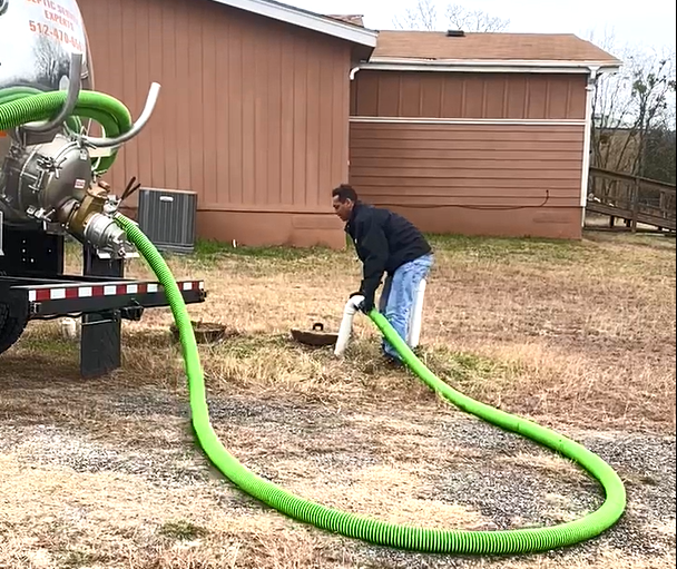 Man connecting green hose to septic tank truck, brown building in background.