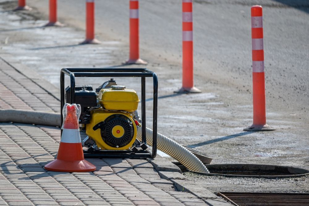 A gasoline water pump sits on a sidewalk near an open manhole, with orange safety bollards and a cone.