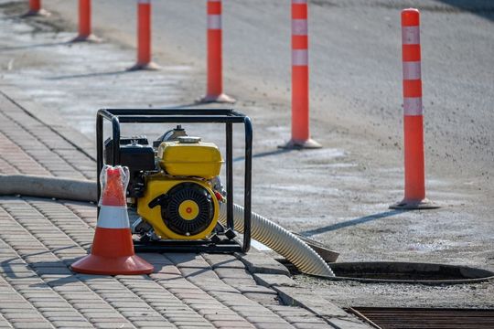 A gasoline water pump sits on a sidewalk near an open manhole, with orange safety bollards and a cone.