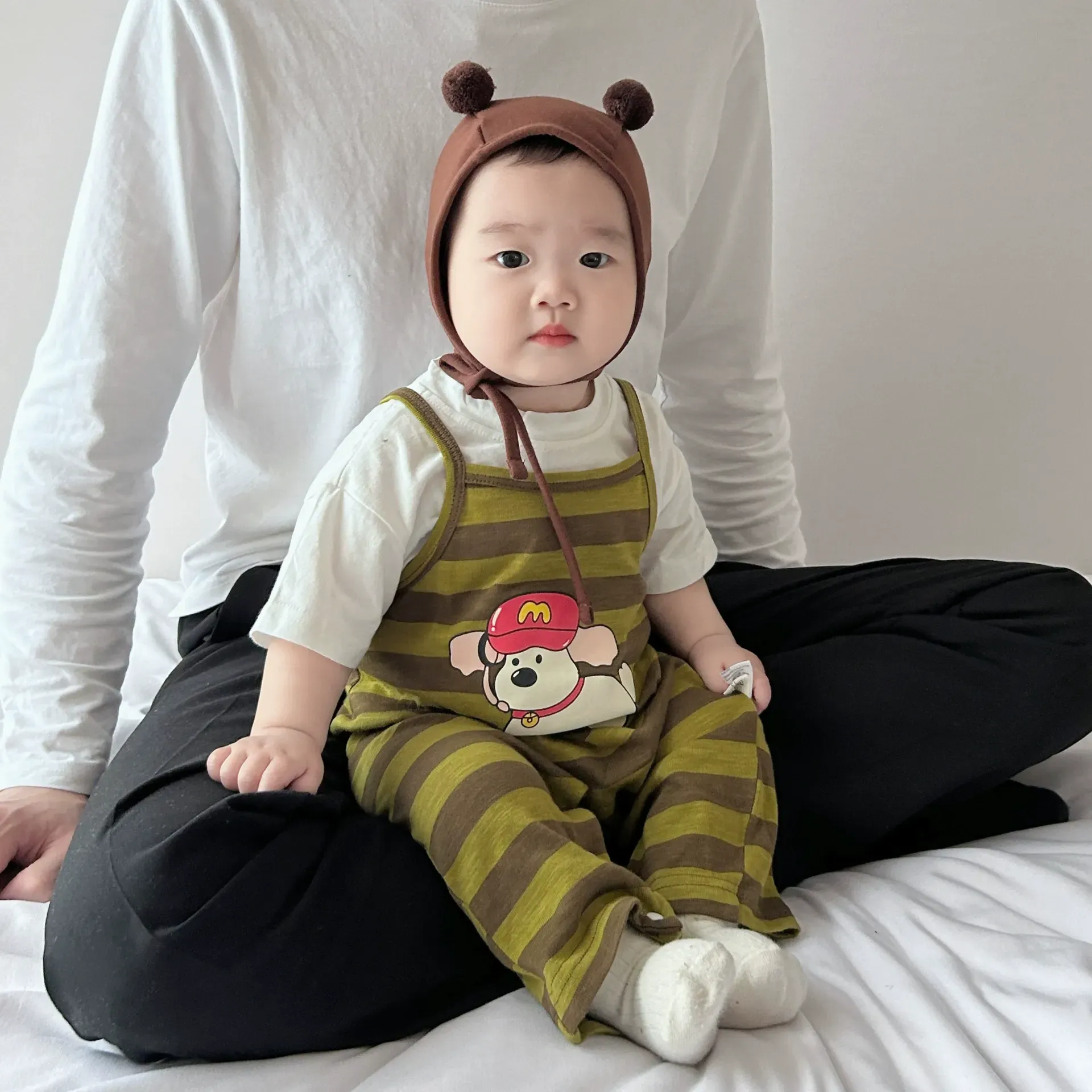 一名戴著帽子、穿著工作服的嬰兒坐在床上，旁邊是一名男子。