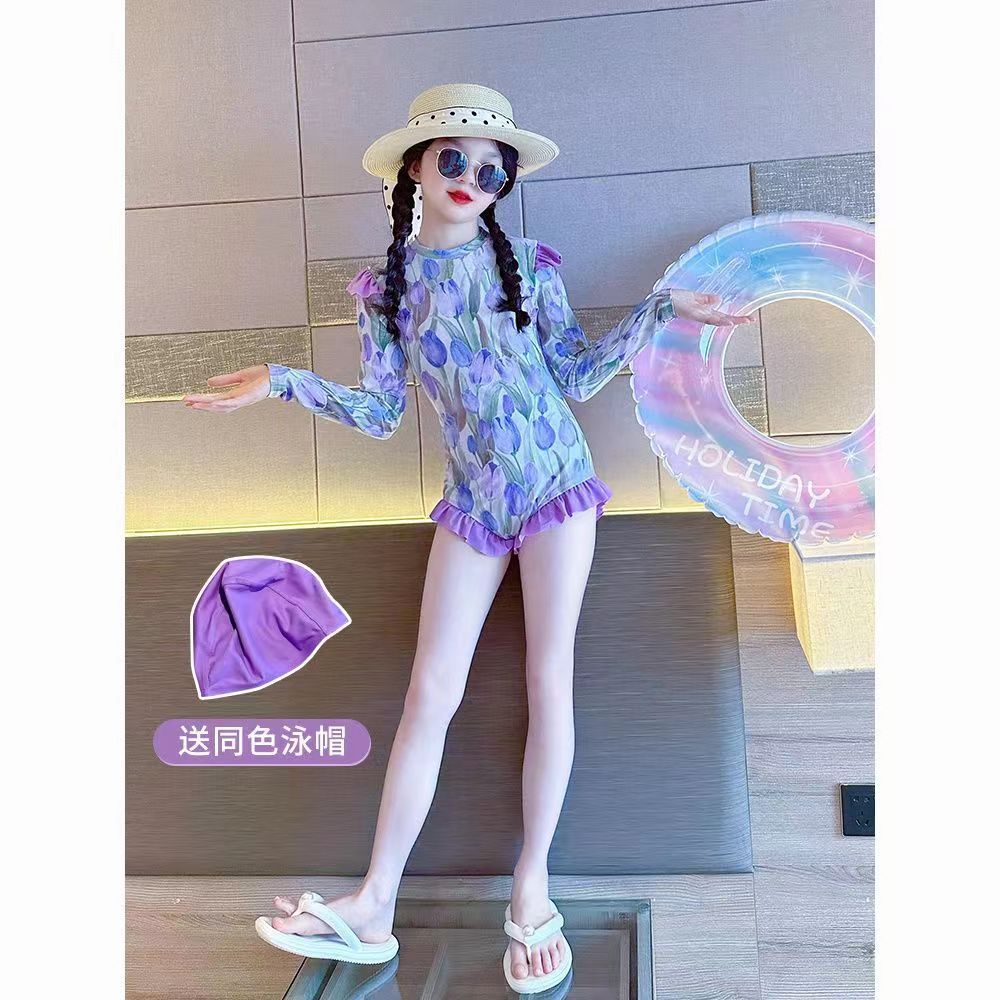 「穿著紫色印花連身泳衣的女孩擺出時尚姿勢，搭配草帽與墨鏡，背景有彩色游泳圈，並附贈同色泳帽。」