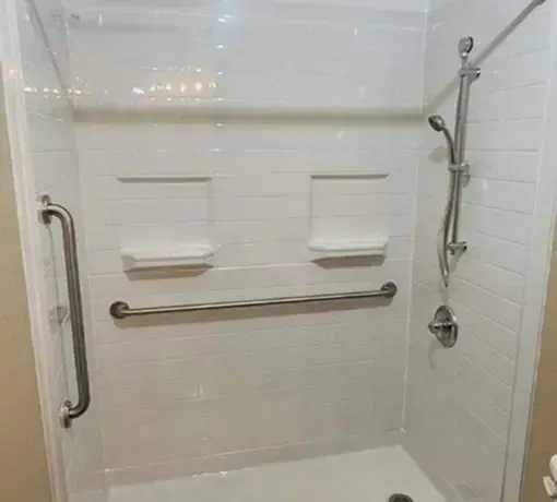 A white, tiled shower stall equipped with a vertical grab bar, a horizontal safety bar, and a handheld shower head.