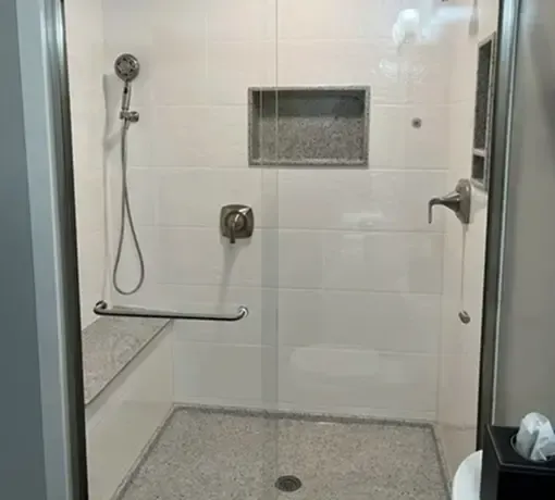 A walk-in shower with white tiled walls, a glass door, a built-in bench, a silver handheld showerhead, and a niche.