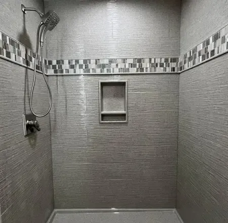 A grey tiled shower stall featuring a chrome handheld shower head, a built-in recessed shelf, and a decorative tile border.
