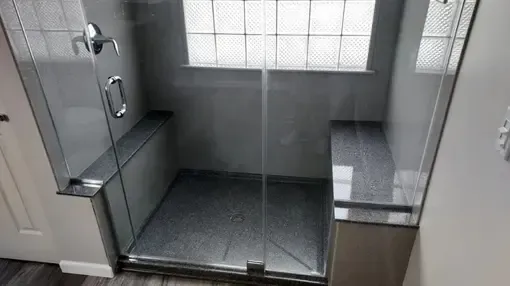 A walk-in shower with gray tile, glass walls, a bench on each side, and a glass block window.