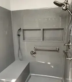 A grey shower stall with a built-in seat, a handheld showerhead, two recessed storage niches, and a metal grab bar.