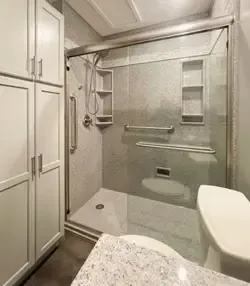 A modern walk-in shower with a glass enclosure, grab bars, built-in shelving, and white cabinets in a bright bathroom.