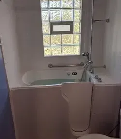 A white walk-in bathtub with a side-entry door, a grab bar, and a handheld shower head set beneath a glass block window.