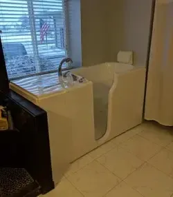A white walk-in bathtub with a side door and chrome faucet installed in a tiled bathroom with a window and blinds.