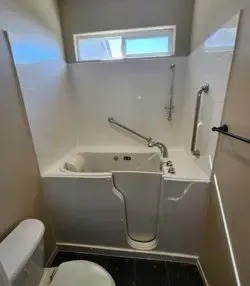 A walk-in bathtub with a white frame and safety grab bars, located in a bathroom with a window and a nearby toilet.