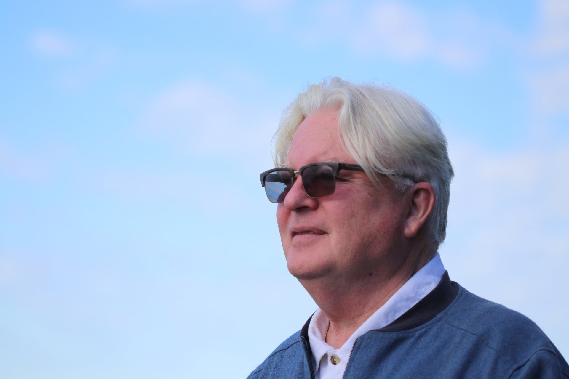A man wearing sunglasses and a blue jacket is standing in front of a blue sky.