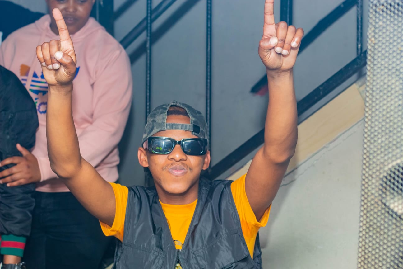A young man wearing sunglasses and a hat is raising his arms in the air.