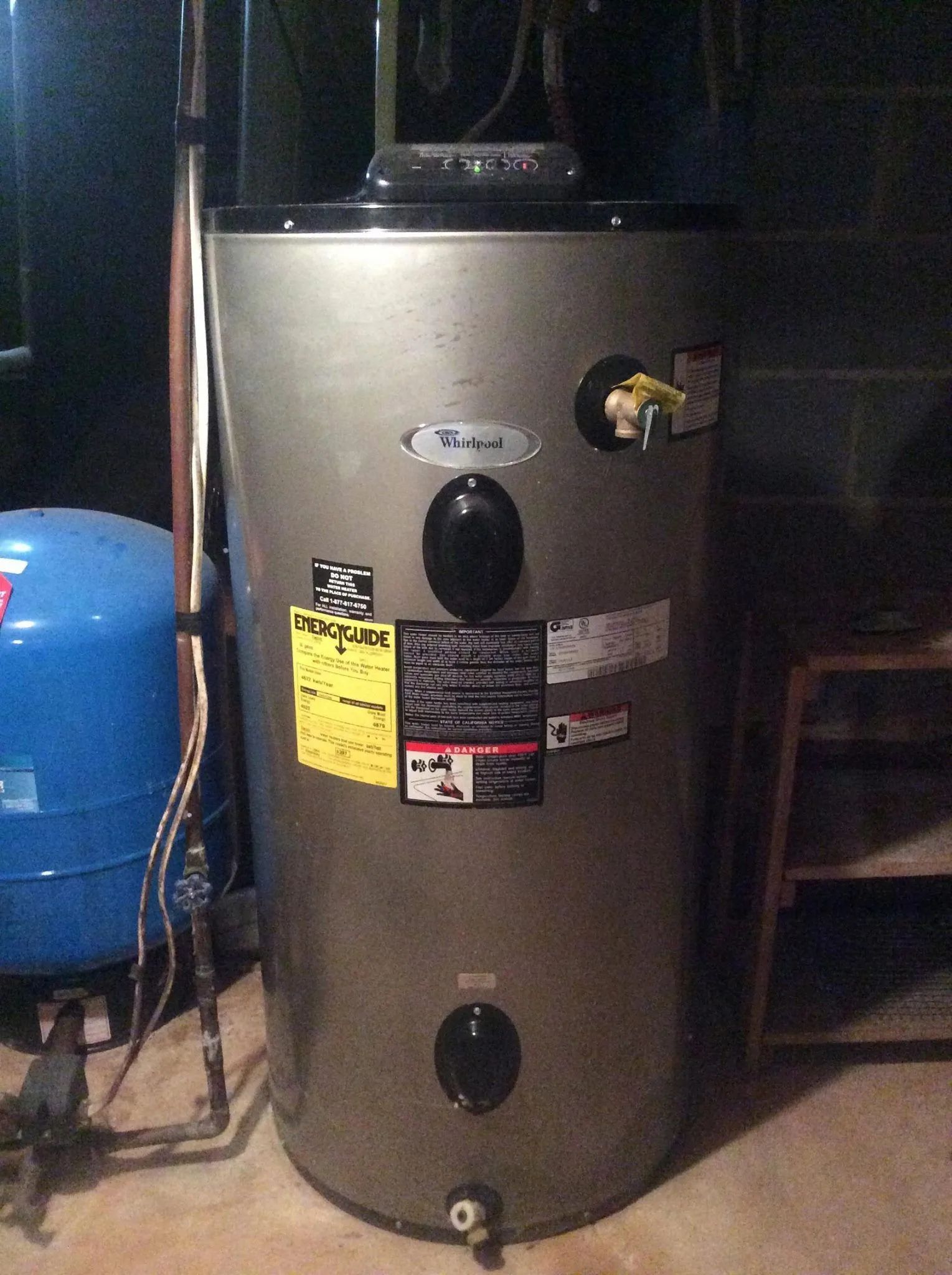 A gray cylindrical electric water heater standing in a utility area next to a blue pressure tank.