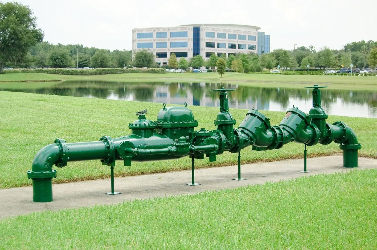 Green industrial pipes on a grassy path by a lake, modern building in the background.