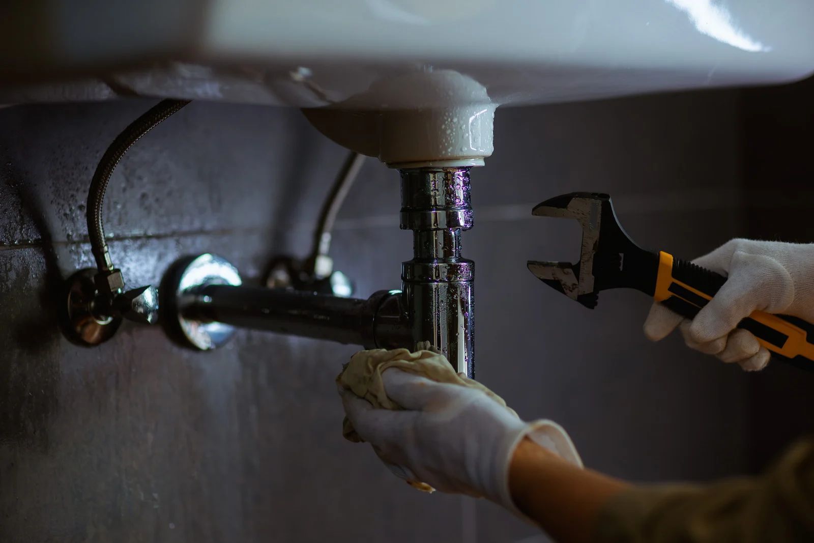 Plumber in gloves fixing a leaky bathroom sink with a wrench.