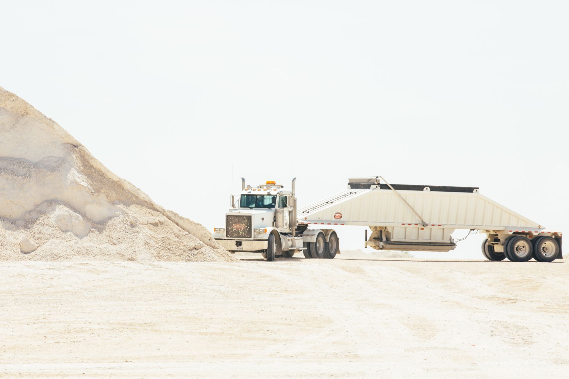 A white semi truck is driving down a dirt road next to a pile of sand.