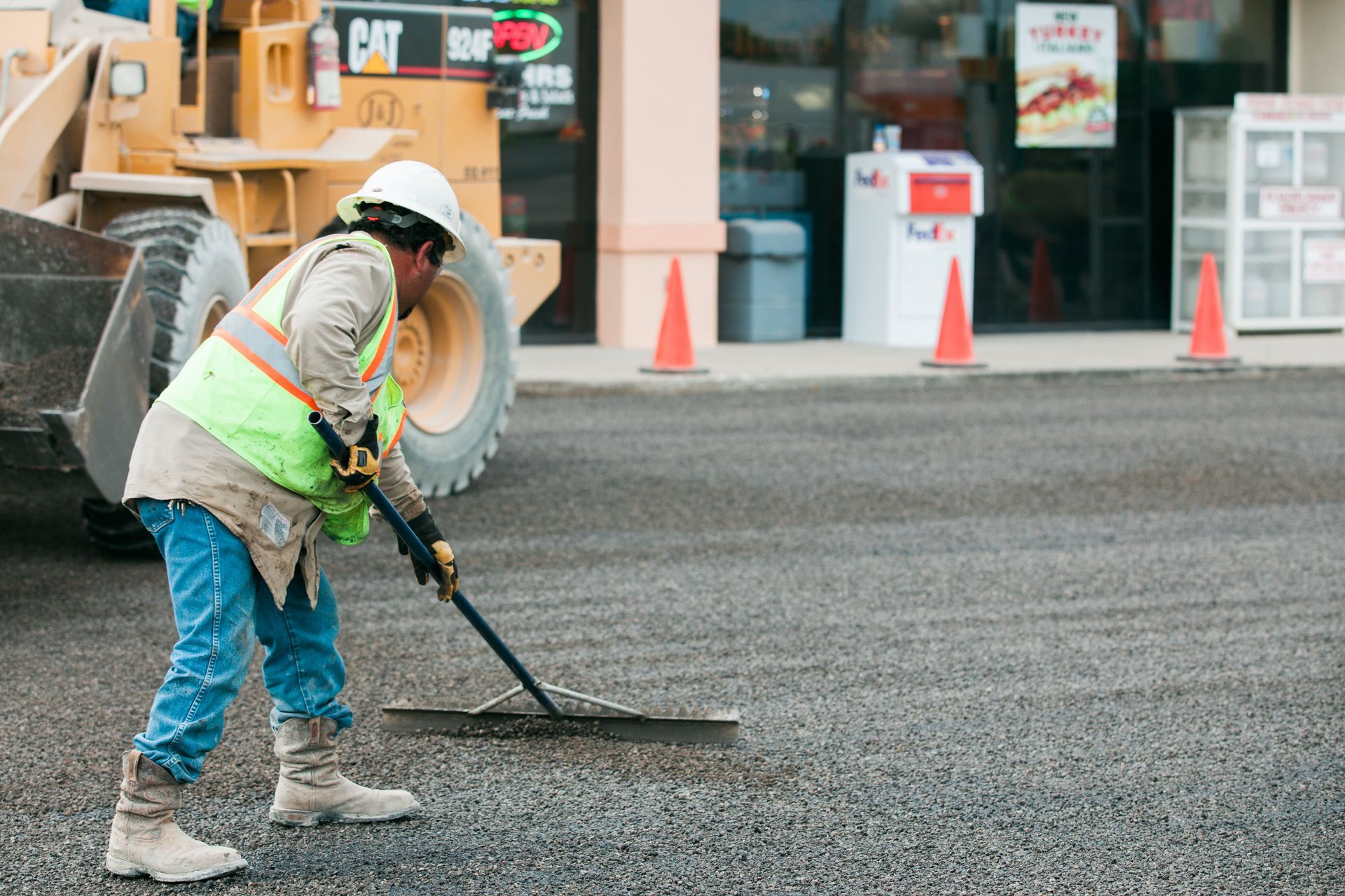 A woman wearing a safety vest and hard hat is using a rake to clean a parking lot.
