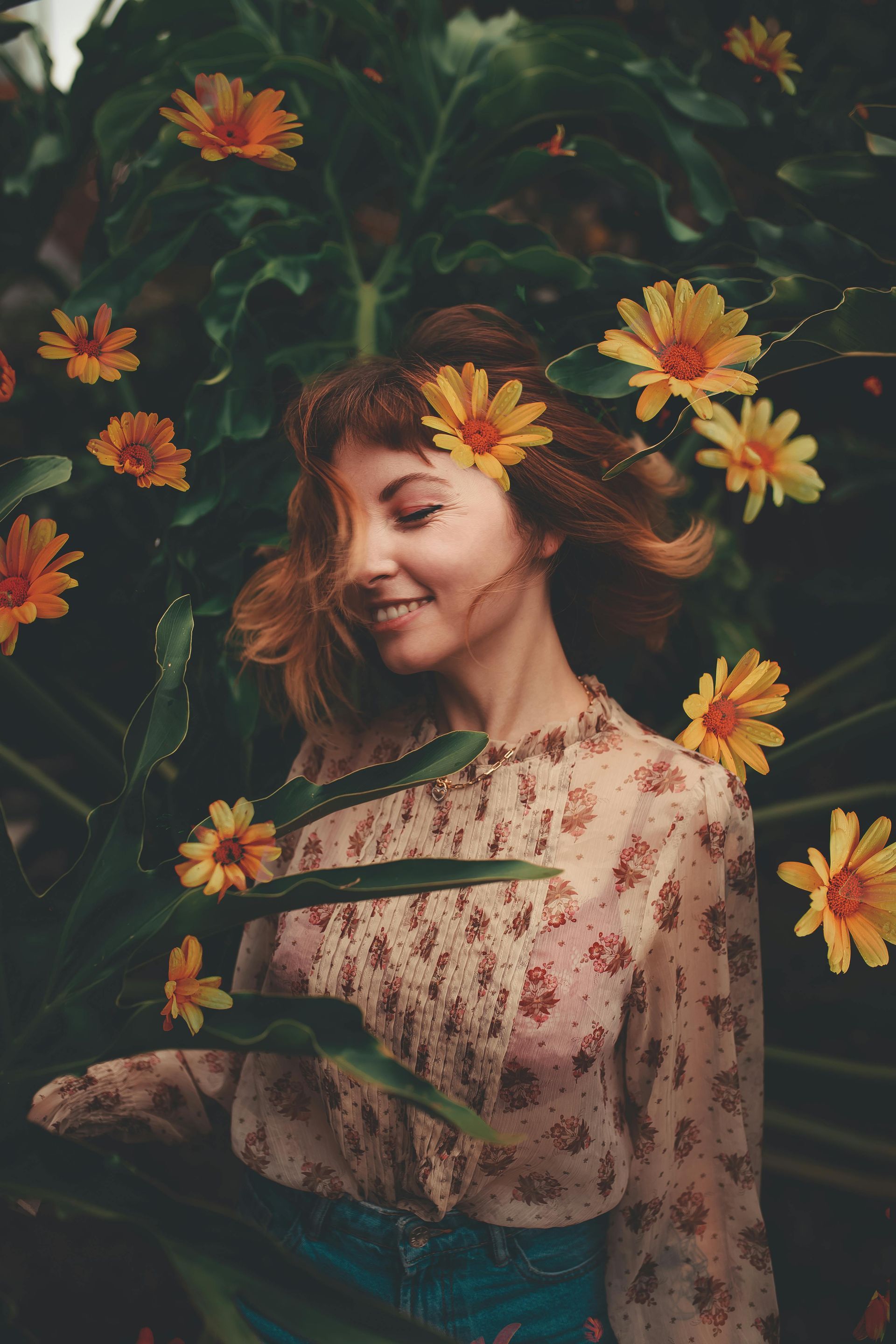 Woman with auburn hair smiles amidst yellow flowers and green foliage. She wears a floral blouse.