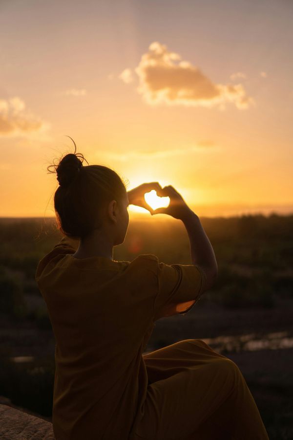 Silhouette of person making heart shape with hands, sunset through heart.