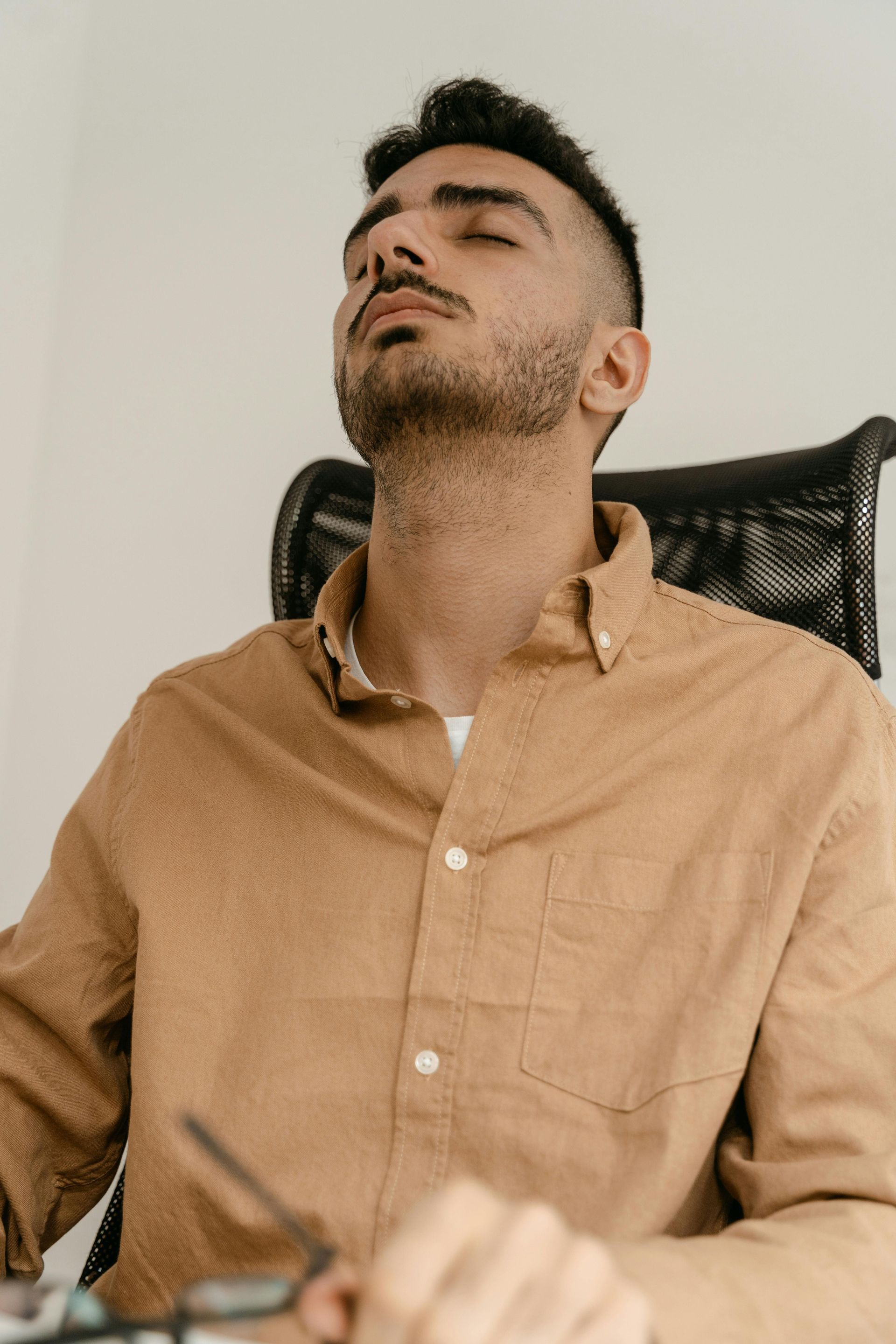 Man sitting at a desk with eyes closed, leaning back with a neutral expression. Brown shirt.