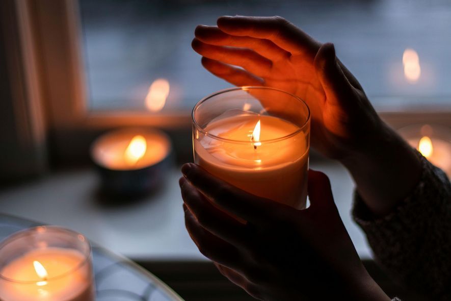 Hands cupping a lit candle in a glass holder, soft orange glow. Other candles in the background.