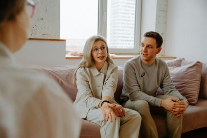 Couple in therapy session, sitting on pink couch, talking to therapist.