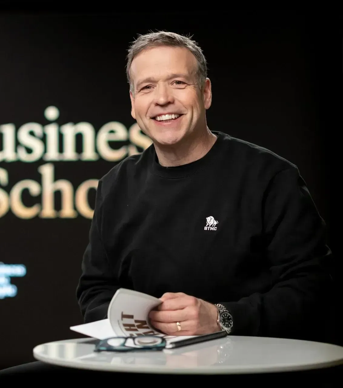 A man is sitting at a table in front of a sign that says business school