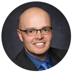 A bald man wearing glasses and a blue shirt and tie is smiling in a circle.
