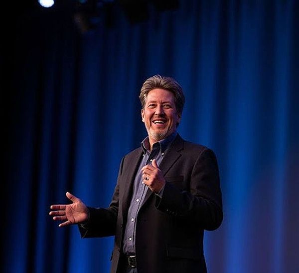 A man in a suit is giving a speech on a stage in front of a blue curtain.