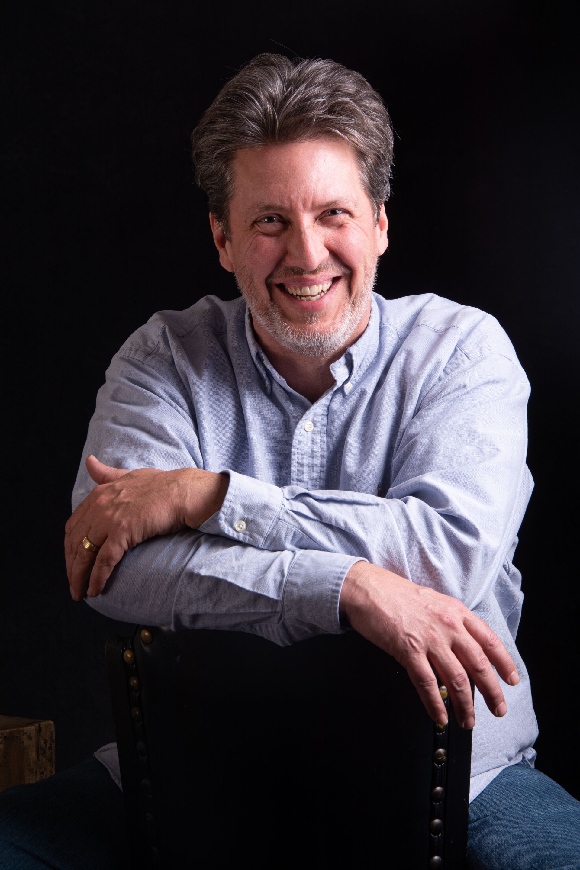 A man with a beard is sitting in a chair with his arms crossed and smiling.