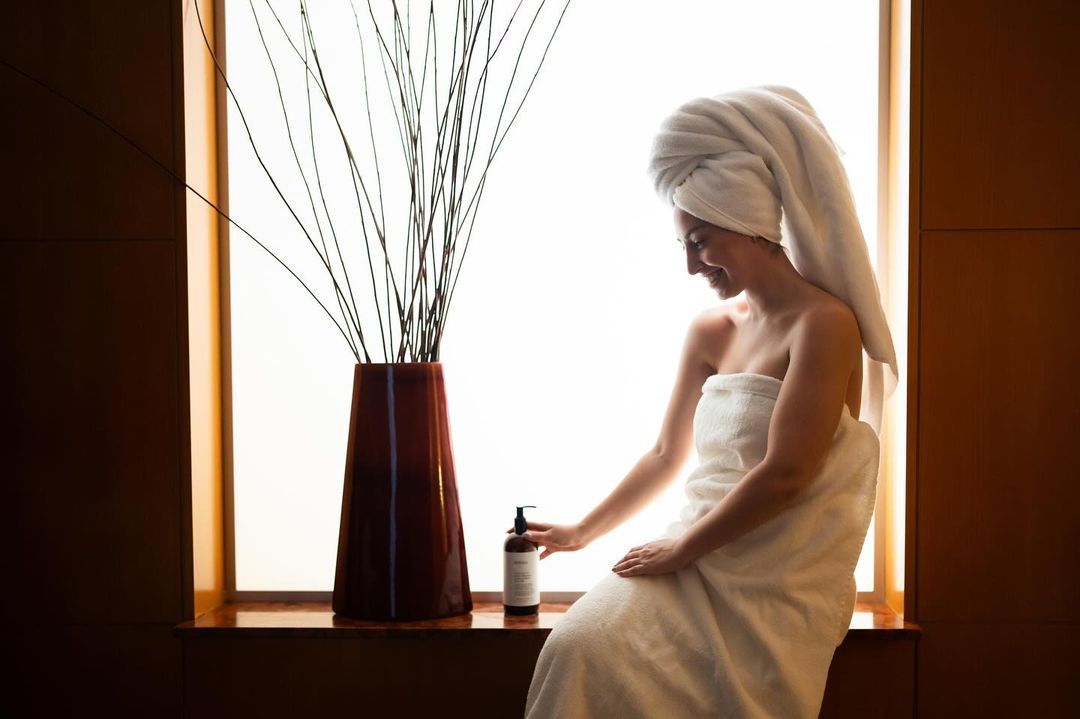 Una mujer envuelta en una toalla está sentada en el alféizar de una ventana sosteniendo una botella de loción.