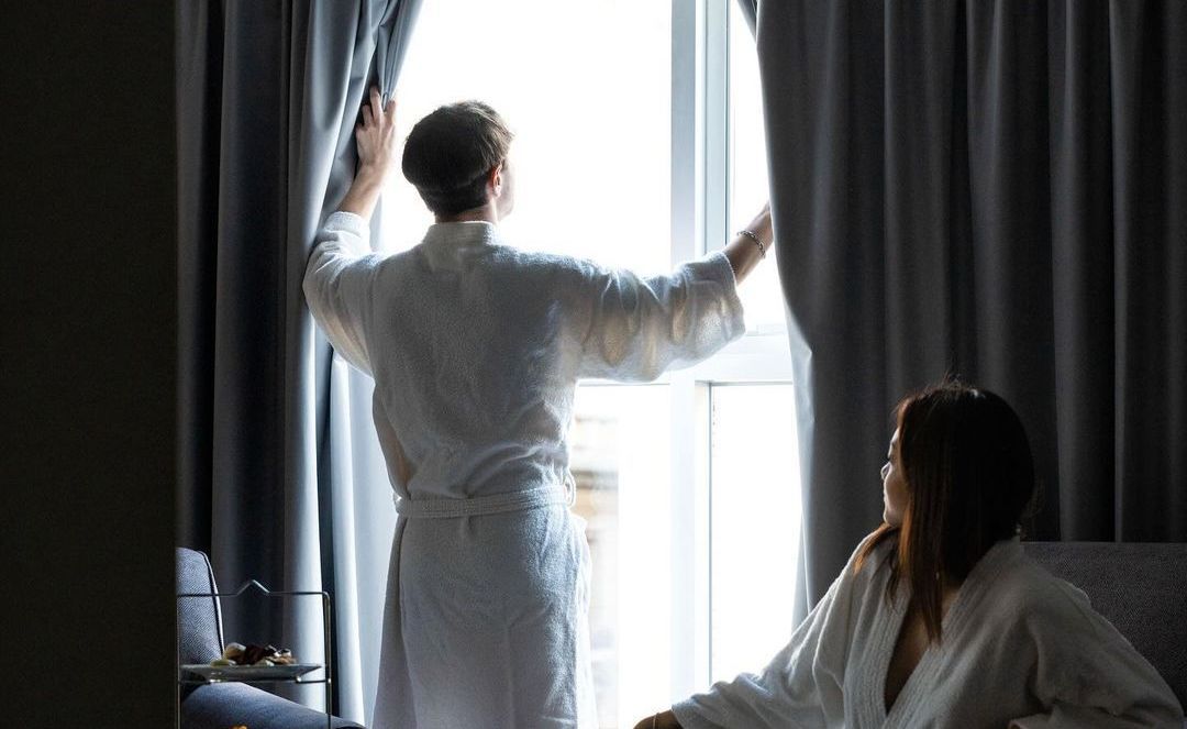 Un hombre y una mujer abren las cortinas de una habitación de hotel.