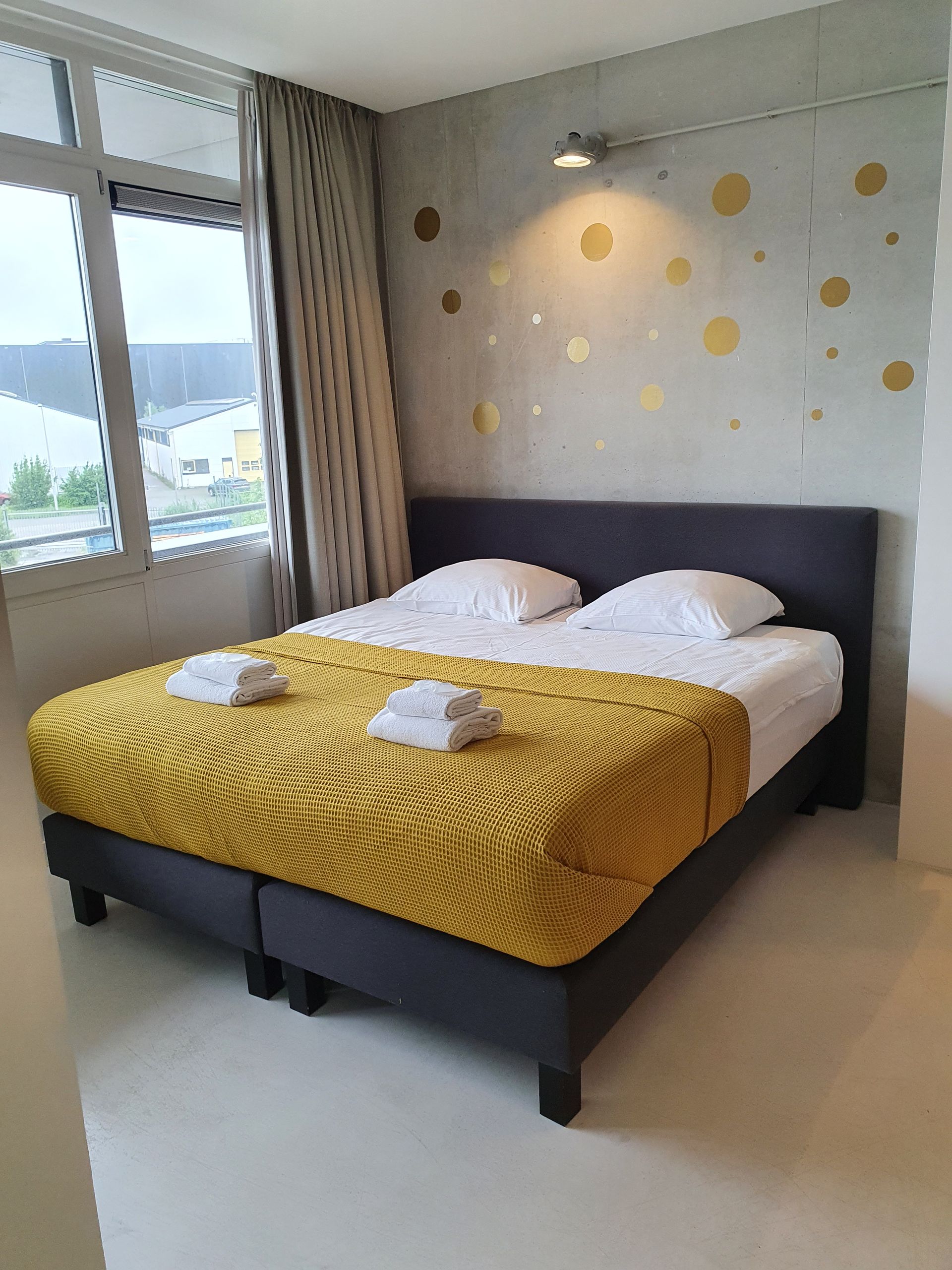 Slaapkamer met een grijs bedframe en een gele deken. Wit beddengoed en gouden stippen op de grijze muur.