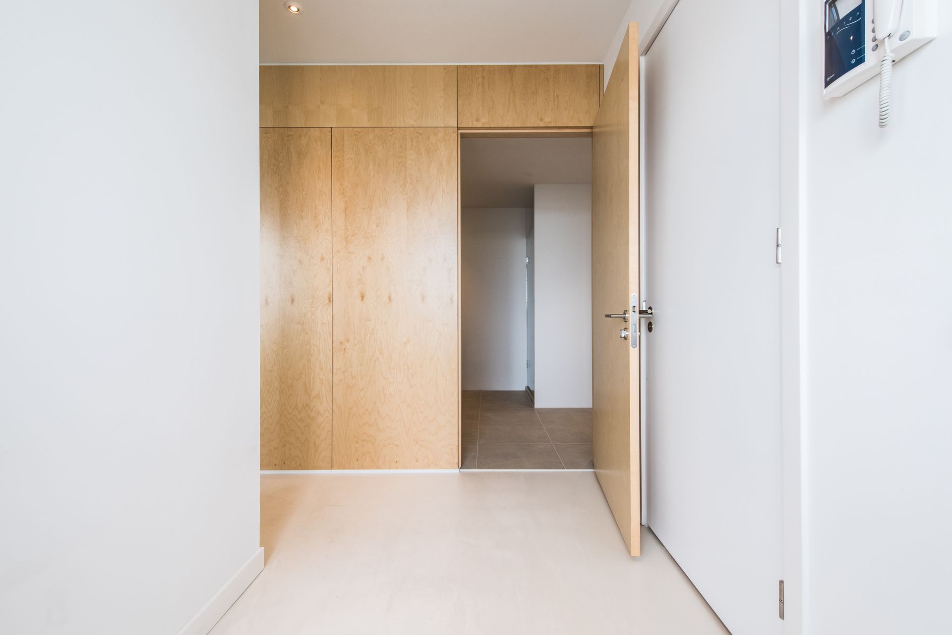 Hal met witte muren en vloer, houten opbergkast en open deur.