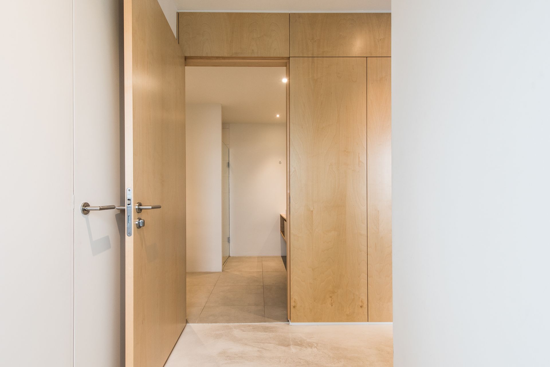 Een lichte houten deur opent zich en geeft toegang tot een hal met lichte muren, vloeren en een deur aan het einde.