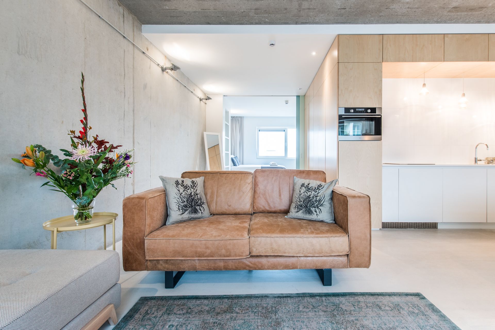 Woonkamer met een bruine leren bank, bloemen en een vloerkleed. Lichte houten kasten en betonnen muren.