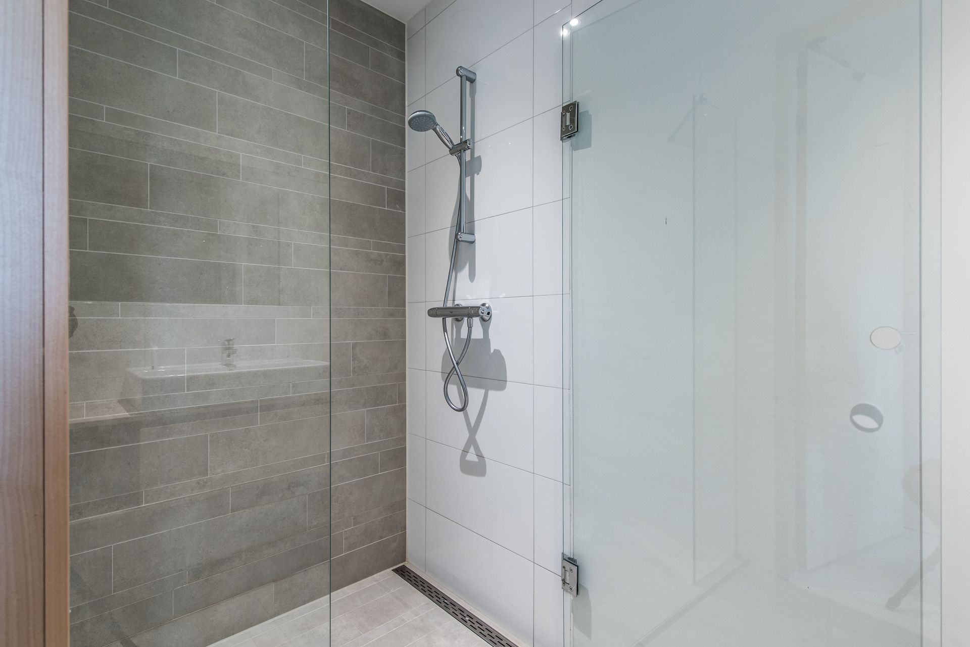 Badkamer met een glazen douchecabine. Grijze tegelwand met douchekop en witte muren.