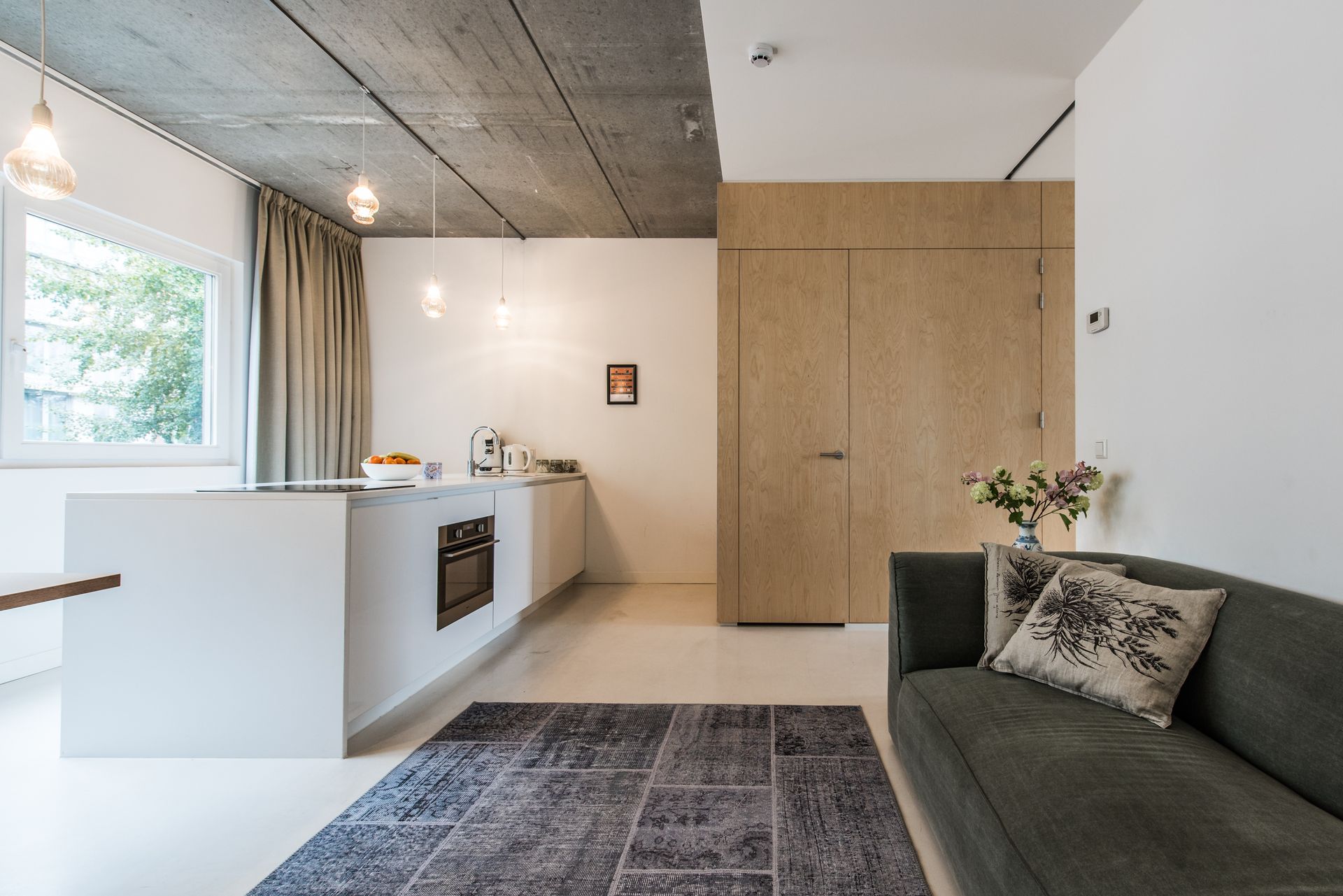 Moderne, minimalistische keuken met kookeiland, houten deur en grijze bank, onder een betonnen plafond.