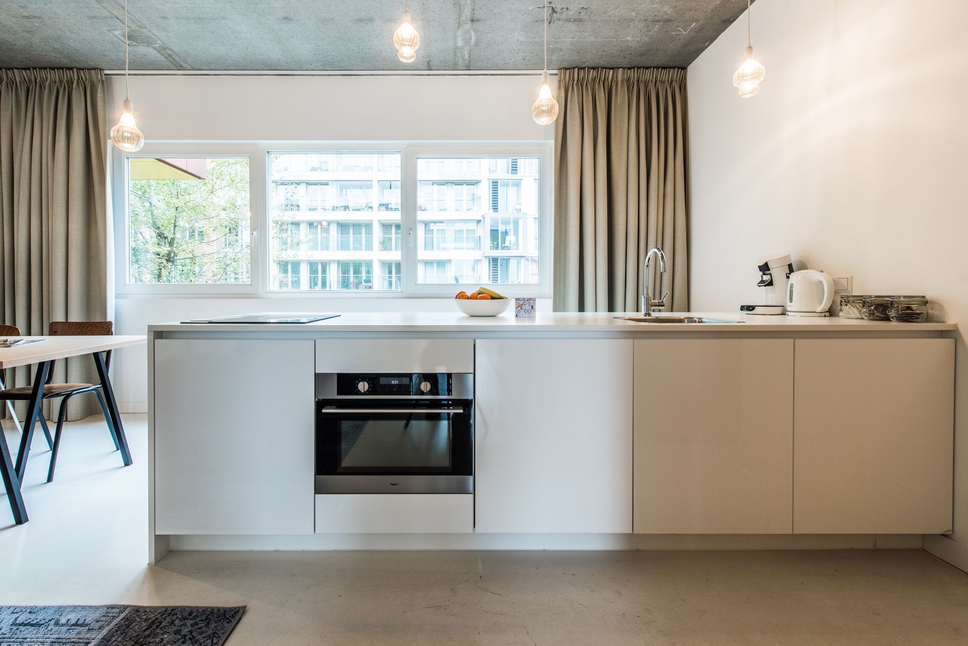 Moderne witte keuken met ingebouwde oven, groot raam, hanglampen en beige gordijnen.