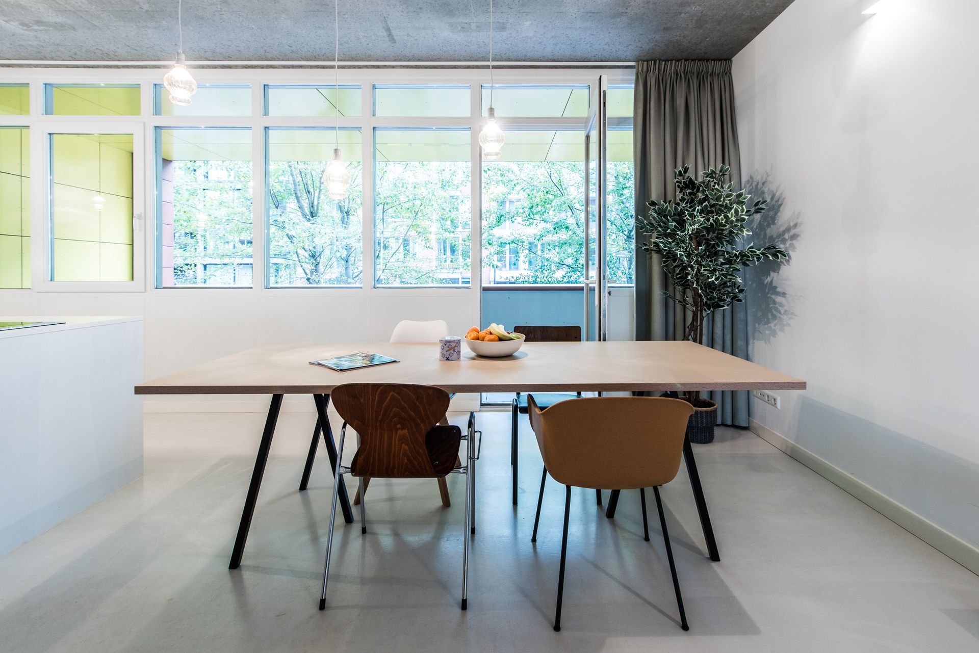 Eetkamer met een lichte houten tafel, metalen poten en kleurrijke stoelen. Groot raam en een boom met veel bladeren.