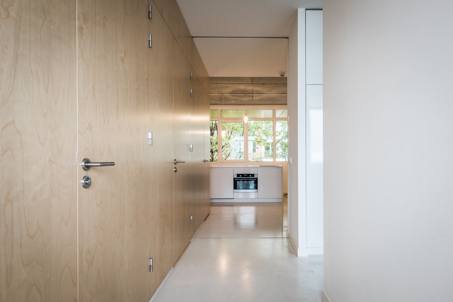 Gang met aan één zijde lichte houten lambrisering, die toegang geeft tot een lichte, witte keuken met een raam.