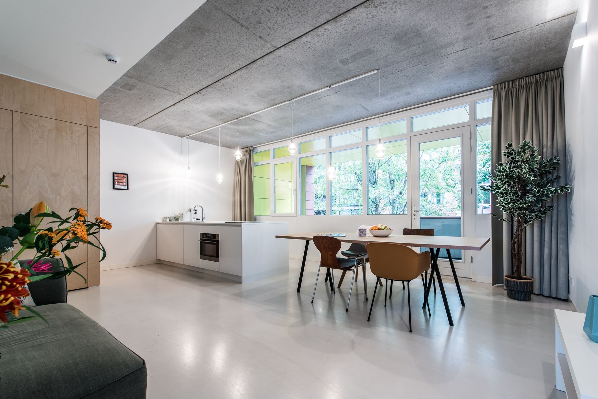 Moderne woonruimte met betonnen plafond, witte muren en grote ramen met uitzicht op bomen.